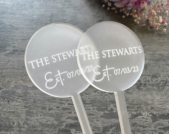 Puede incluir: Dos letreros redondos de acrílico transparente con texto blanco. Los letreros dicen "The Stewarts Est. 07/03/23" y "The Stewarts Est. 07/0/0". Los letreros están unidos a palos de acrílico transparente.