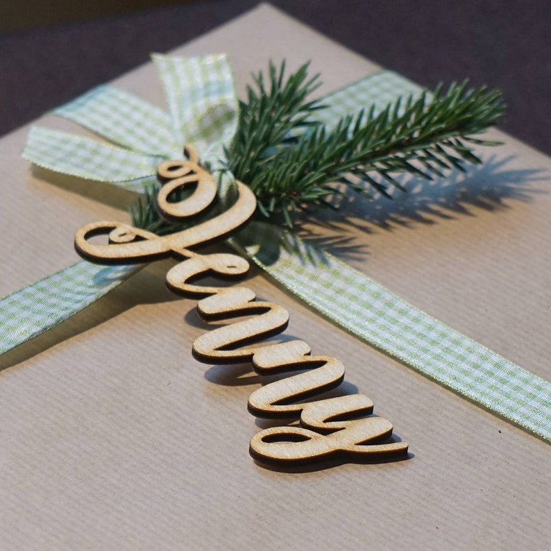 Puede incluir: Una cinta de gingham verde claro y blanco atada en un lazo con una etiqueta de nombre de madera que dice "Jenny" en una caja de regalo marr&oacute;n.