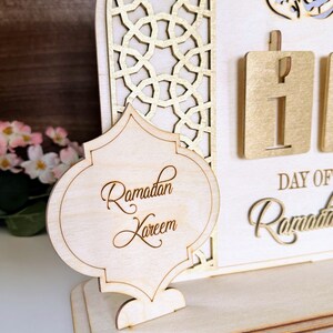 May include: A wooden Ramadan countdown calendar with a gold-painted intricate design. The calendar features a sign that says "Ramadan Kareem" and a number display for the day of Ramadan.