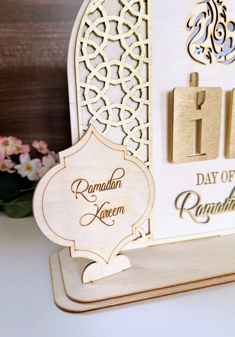 May include: A wooden Ramadan countdown calendar with a gold-painted intricate design. The calendar features a sign that says "Ramadan Kareem" and a number display for the day of Ramadan.