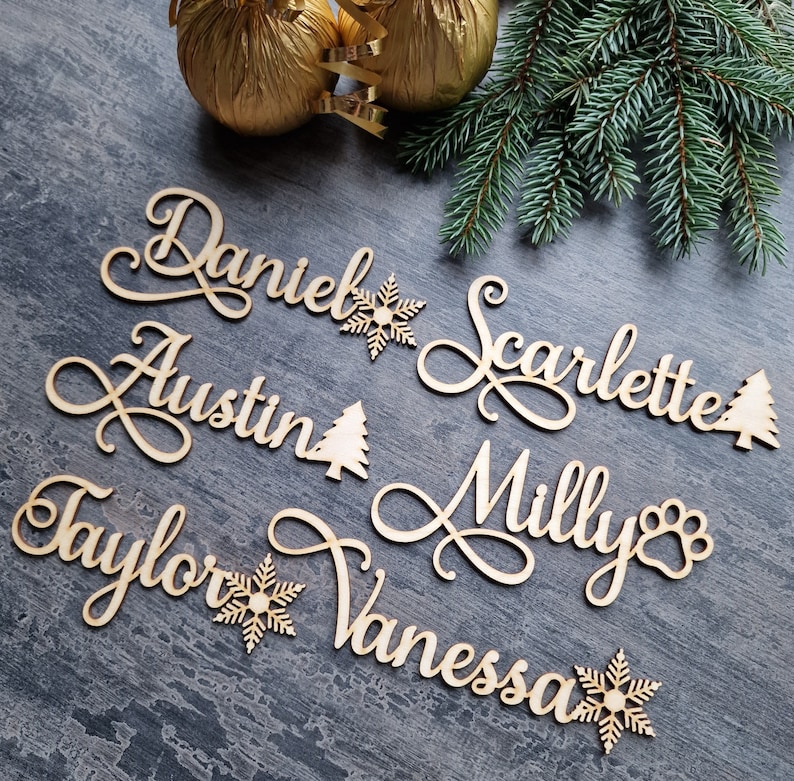May include: Wooden Christmas ornaments with names "Daniel", "Austin", "Scarlette", "Milly", "Taylor", and "Vanessa" in cursive script. Each name is accompanied by a snowflake or a Christmas tree.