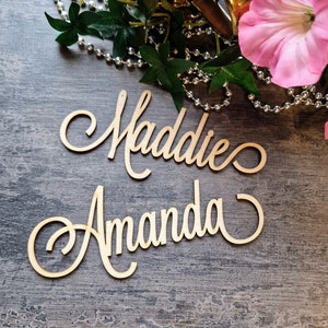 May include: Two wooden signs with the names Maddie and Amanda written in cursive script. The signs are on a gray textured surface.