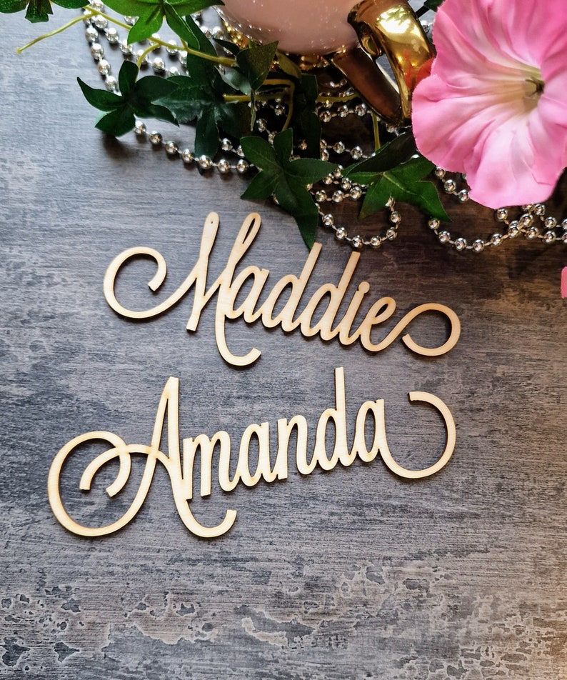 May include: Two wooden signs with the names Maddie and Amanda written in cursive script. The signs are on a gray textured surface.