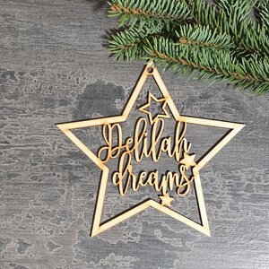 Puede incluir: Adorno de madera en forma de estrella con las palabras "Delilah dreams" recortadas en una fuente cursiva. El adorno tiene un peque&ntilde;o agujero para colgar y est&aacute; rodeado por una forma de estrella m&aacute;s grande. Una ramita de hoja perenne est&aacute; en el fondo.