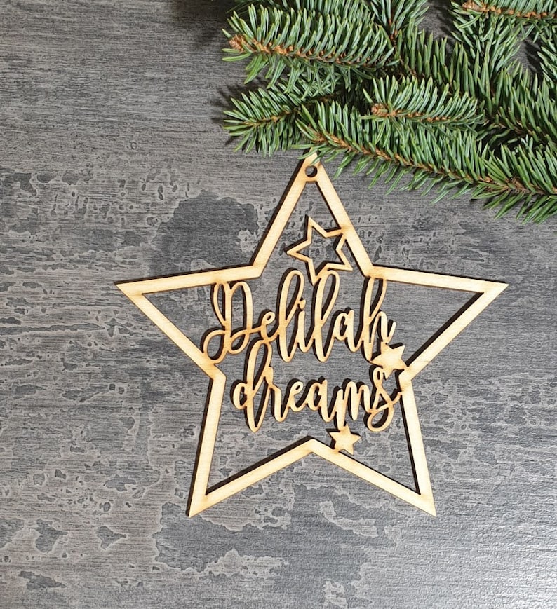 Puede incluir: Adorno de madera en forma de estrella con las palabras "Delilah dreams" recortadas en una fuente cursiva. El adorno tiene un peque&ntilde;o agujero para colgar y est&aacute; rodeado por una forma de estrella m&aacute;s grande. Una ramita de hoja perenne est&aacute; en el fondo.