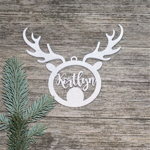 May include: A silver glitter reindeer head ornament with the name "Kortlyn" cut out in the center. The ornament has antlers and a circular face. A sprig of blue-green pine needles is in the corner.