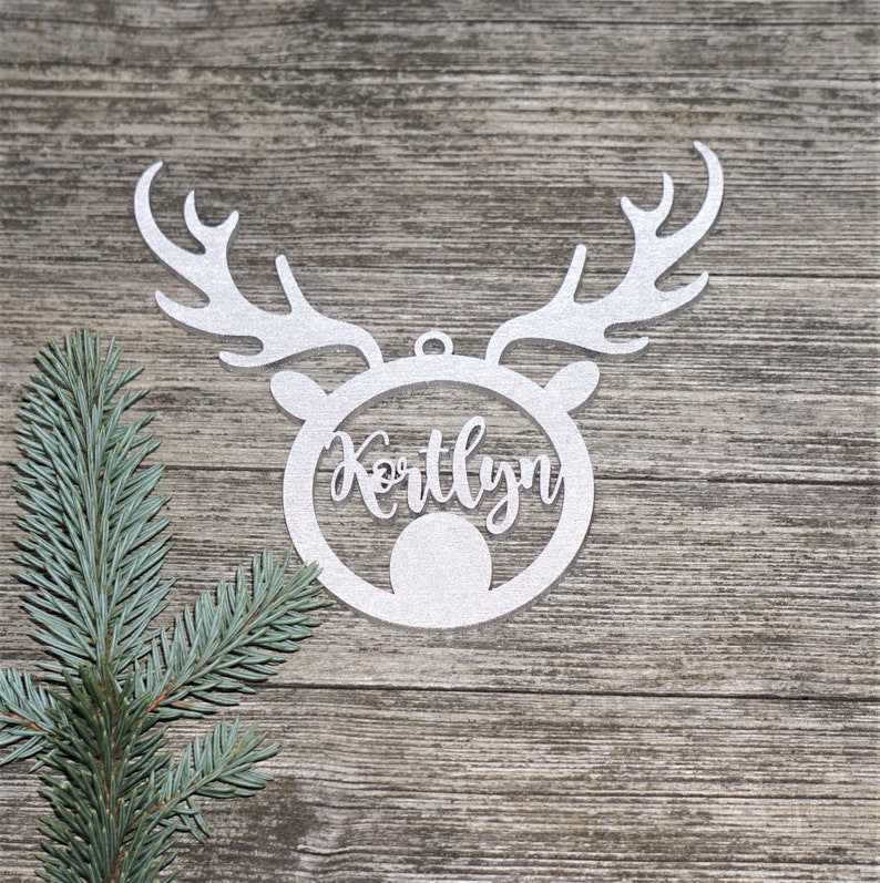 May include: A silver glitter reindeer head ornament with the name "Kortlyn" cut out in the center. The ornament has antlers and a circular face. A sprig of blue-green pine needles is in the corner.