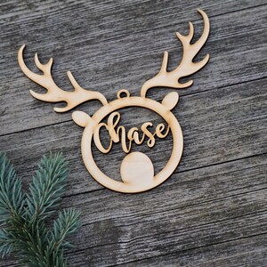 May include: A wooden Christmas ornament in the shape of a reindeer head with antlers. The name "Chase" is cut out in a cursive font within the reindeer's face. A sprig of blue-green pine needles is in the lower left corner.
