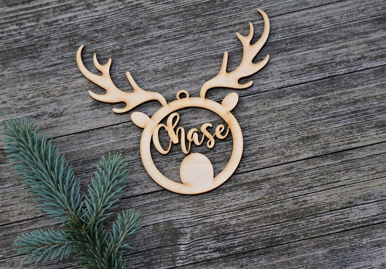 May include: A wooden Christmas ornament in the shape of a reindeer head with antlers. The name "Chase" is cut out in a cursive font within the reindeer's face. A sprig of blue-green pine needles is in the lower left corner.