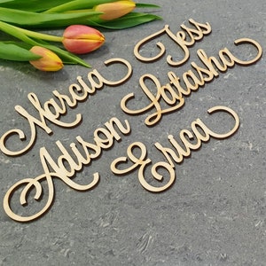 May include: Wooden cutout names in cursive script, including Marcia, Addison, Jess, Latasha, and Erica. The names are arranged in a semi-circle on a gray background with yellow and pink flowers.