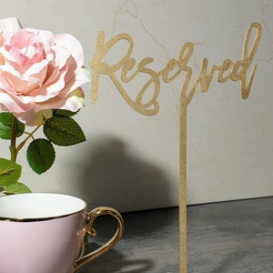 May include: Gold glitter table sign with the word "Reserved" in a cursive font. The sign is on a gold stand with a cross base.