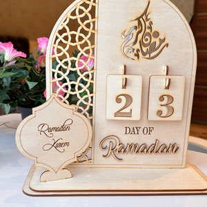 May include: A wooden Ramadan calendar with a cutout design of an archway and Arabic calligraphy. The calendar features two flippable number tiles displaying the number 23, with the text "Day of Ramadan" below. The calendar also includes a separate wooden plaque with the text "Ramadan Kareem".