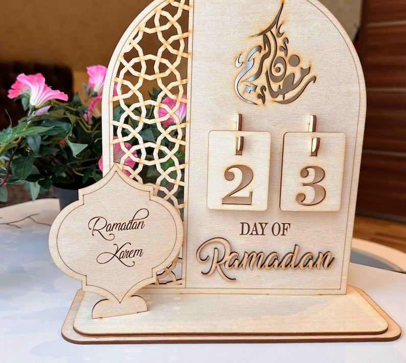 May include: A wooden Ramadan calendar with a cutout design of an archway and Arabic calligraphy. The calendar features two flippable number tiles displaying the number 23, with the text "Day of Ramadan" below. The calendar also includes a separate wooden plaque with the text "Ramadan Kareem".