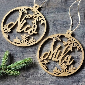 May include: Two wooden Christmas ornaments with snowflake designs. The ornaments are brown and have the names "Nico" and "Mila" written on them.