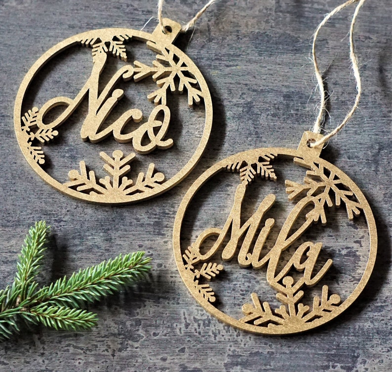May include: Two wooden Christmas ornaments with snowflake designs. The ornaments are brown and have the names "Nico" and "Mila" written on them.