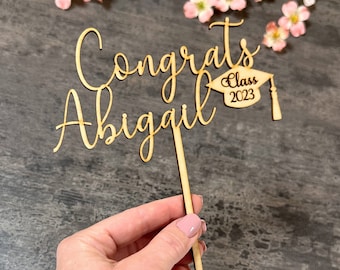 May include: Wooden cake topper with the text "Congrats Abigail" and a graduation cap with the text "Class 2023".