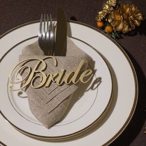 May include: A formal table setting with a gold-rimmed plate, a linen napkin, and silverware. A gold-colored wooden cutout that spells "Bride" rests on the napkin. Decorative elements include small wooden snowflakes and a gold floral accent.
