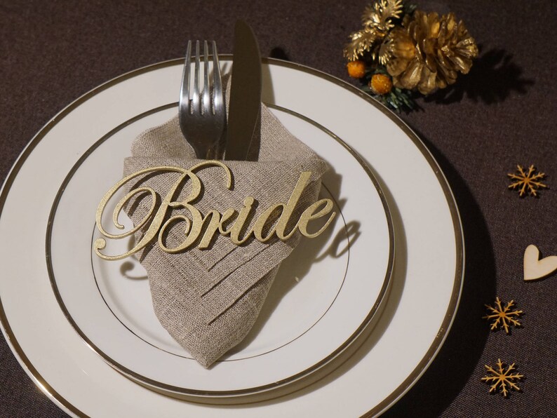 May include: A formal table setting with a gold-rimmed plate, a linen napkin, and silverware. A gold-colored wooden cutout that spells "Bride" rests on the napkin. Decorative elements include small wooden snowflakes and a gold floral accent.