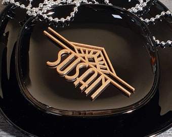 Pode incluir: Uma placa de nome de madeira dourada com o nome "Susan" recortado em um design geométrico. A placa de nome está sobre um prato preto com uma corrente prateada sobre ela.