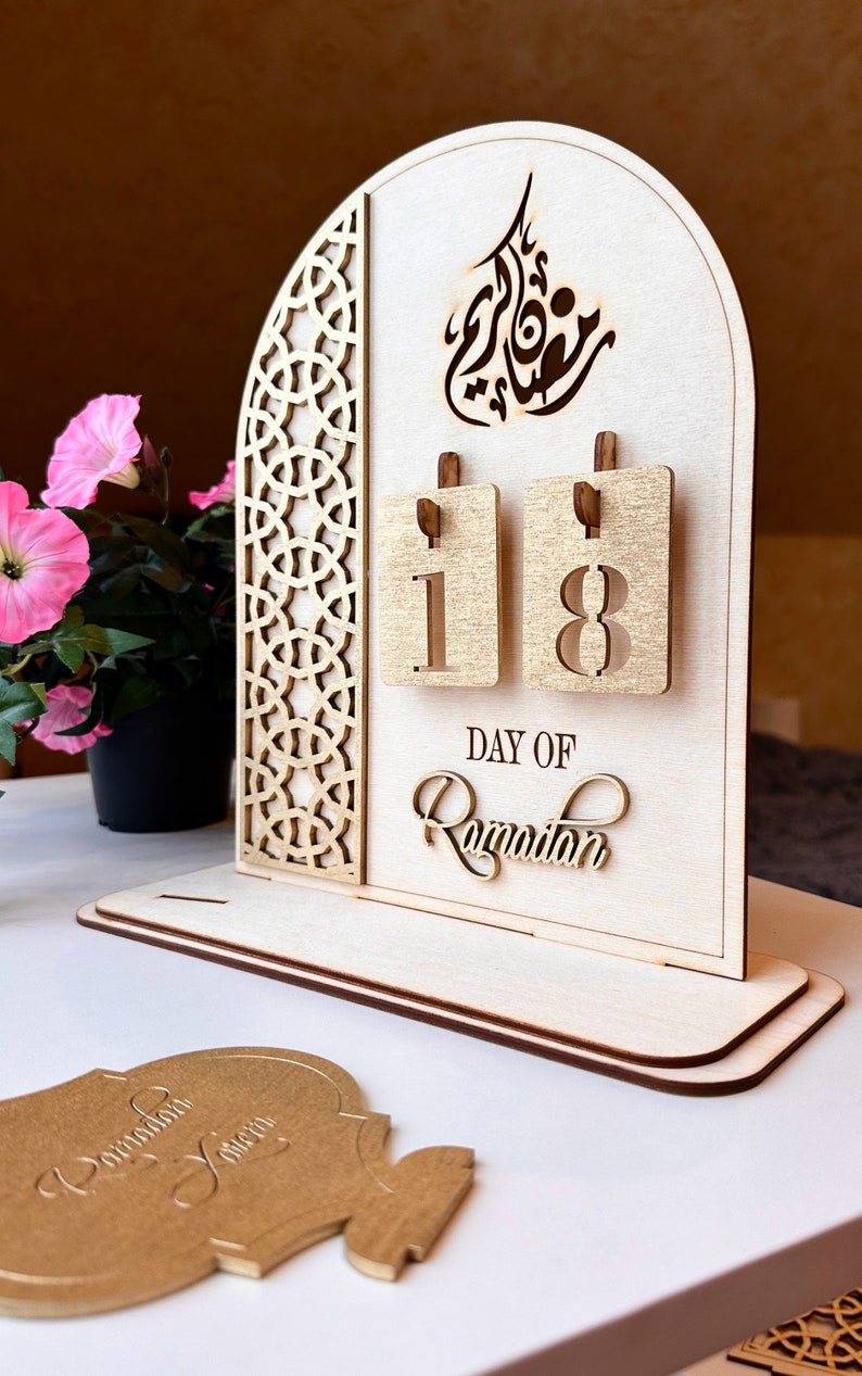 May include: A wooden Ramadan calendar with a decorative arch design and a gold-colored background. The calendar features a flip-style design with two numbers, 1 and 8, indicating the 18th day of Ramadan. The text "DAY OF RAMADAN" is written below the numbers.