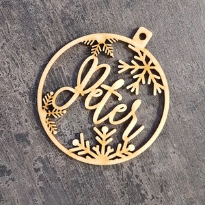 May include: A wooden Christmas bauble with the name "Peter" in cursive script. The bauble is round with snowflake cutouts and a small hole for hanging. The wood has a natural, light brown colour.