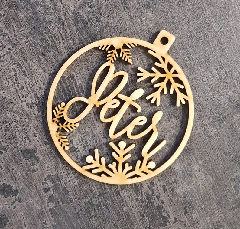 May include: A wooden Christmas bauble with the name "Peter" in cursive script. The bauble is round with snowflake cutouts and a small hole for hanging. The wood has a natural, light brown colour.