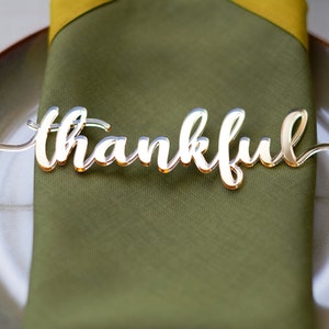 Pode incluir: Um marcador de lugar dourado com a palavra "thankful" em it&aacute;lico est&aacute; sobre um guardanapo verde dobrado em um prato. O marcador de lugar &eacute; feito de um material reflexivo, e o prato tem uma borda dourada. O guardanapo &eacute; verde-oliva.