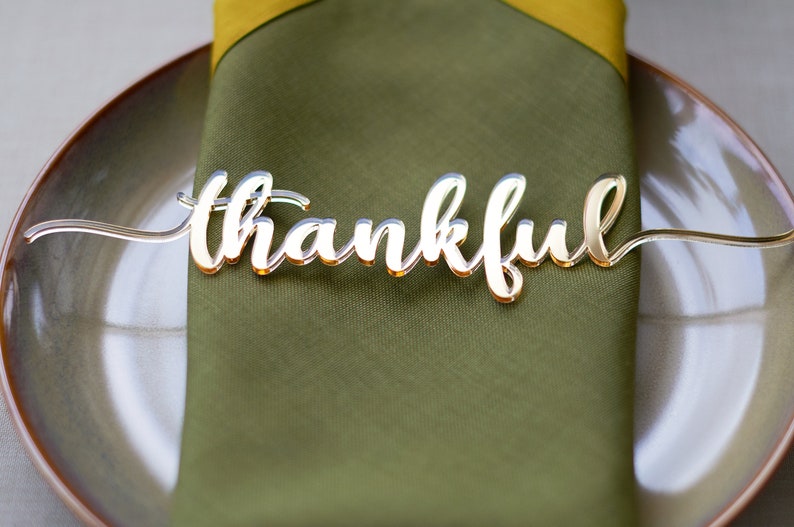Pode incluir: Um marcador de lugar dourado com a palavra "thankful" em it&aacute;lico est&aacute; sobre um guardanapo verde dobrado em um prato. O marcador de lugar &eacute; feito de um material reflexivo, e o prato tem uma borda dourada. O guardanapo &eacute; verde-oliva.