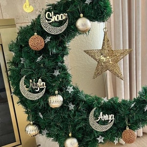 May include: A green crescent moon-shaped wreath decorated with gold stars, gold ornaments, and three gold crescent moons with the names "Shaan", "Niyla", and "Ariya" written on them.
