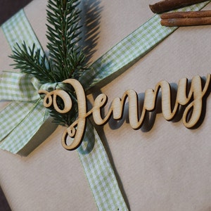 Puede incluir: Una caja de regalo marr&oacute;n con una cinta de cuadros verdes y blancos y una etiqueta de madera que dice "Jenny".
