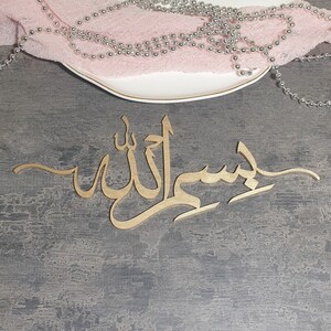Op de afbeelding: Gouden kalligrafie wanddecoratie met het Arabische woord "Insha'Allah" op een grijze achtergrond.