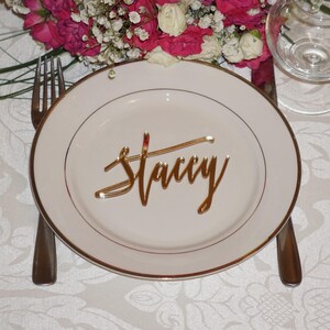 May include: A white plate with a gold rim and a gold name card that says "Stacy" on it. The plate is on a white tablecloth with a floral pattern.