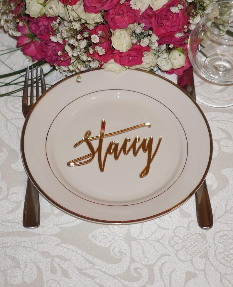 May include: A white plate with a gold rim and a gold name card that says "Stacy" on it. The plate is on a white tablecloth with a floral pattern.