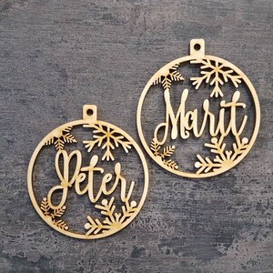 May include: Two wooden Christmas ornaments with snowflake designs. The ornaments are personalized with the names "Peter" and "Marit".
