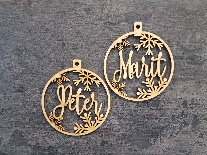 May include: Two wooden Christmas ornaments with snowflake designs. The ornaments are personalized with the names "Peter" and "Marit".