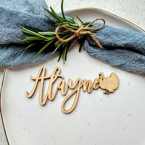 May include: A wooden Thanksgiving place card with the name "Hayme" written in cursive and a small turkey cutout. The place card is on a white plate with a blue napkin and a sprig of rosemary.