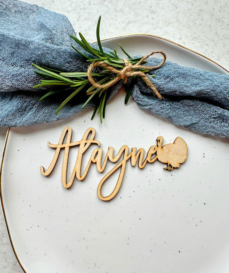 May include: A wooden Thanksgiving place card with the name "Hayme" written in cursive and a small turkey cutout. The place card is on a white plate with a blue napkin and a sprig of rosemary.