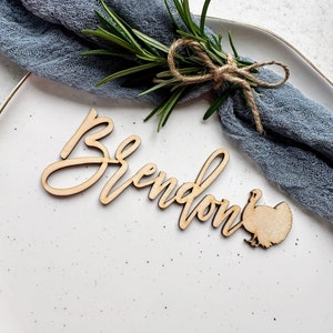 May include: Wooden place card with the name "Brendon" written in cursive and a turkey silhouette. The place card is on a white plate with a blue fabric napkin.