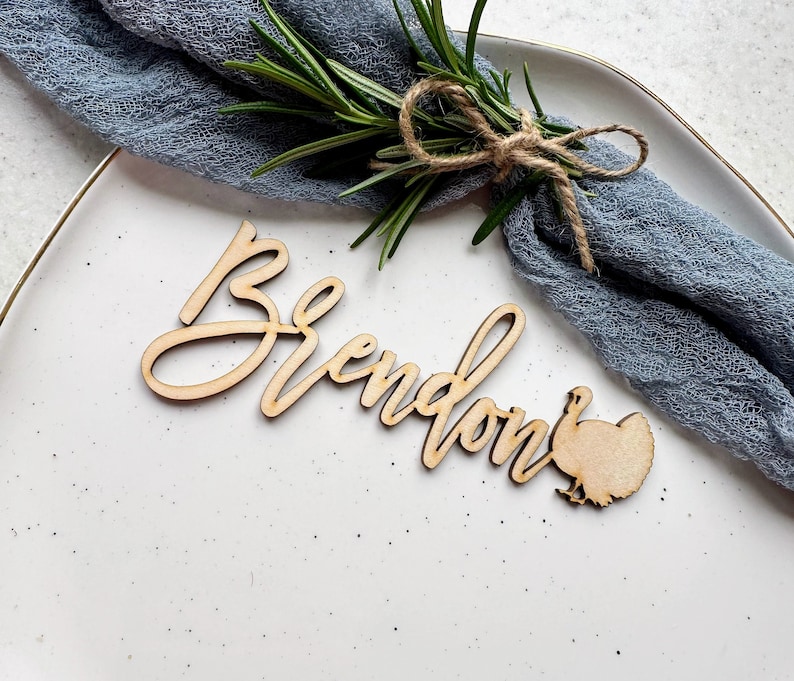 May include: Wooden place card with the name "Brendon" written in cursive and a turkey silhouette. The place card is on a white plate with a blue fabric napkin.