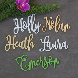 May include: Five wooden name ornaments in different colors. The names are Holly, Nolan, Heath, Laura, and Emerson.