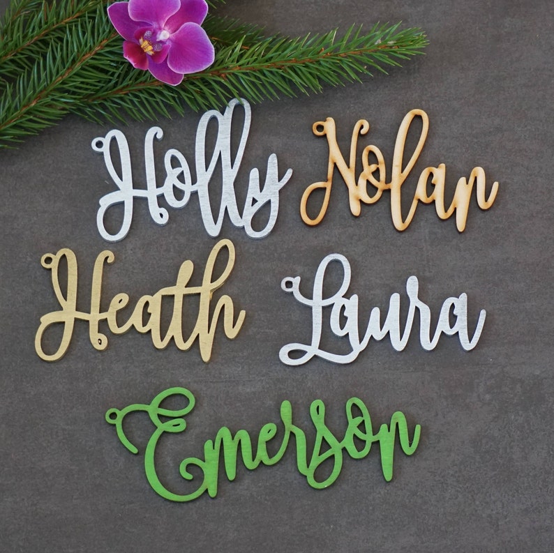 May include: Five wooden name ornaments in different colors. The names are Holly, Nolan, Heath, Laura, and Emerson.