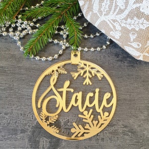May include: A gold Christmas bauble with the name "Stacie" in a cursive font. The round ornament features snowflake cutouts and a hanging loop. Green pine branches and silver beads are in the background.