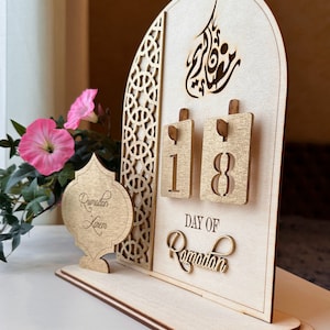 May include: A wooden Ramadan calendar with a gold-painted finish. The calendar features a decorative arch with Arabic calligraphy and a flip-style design to display the day of Ramadan. The calendar is on a wooden base and has a small wooden ornament that says "Ramadan Kareem".