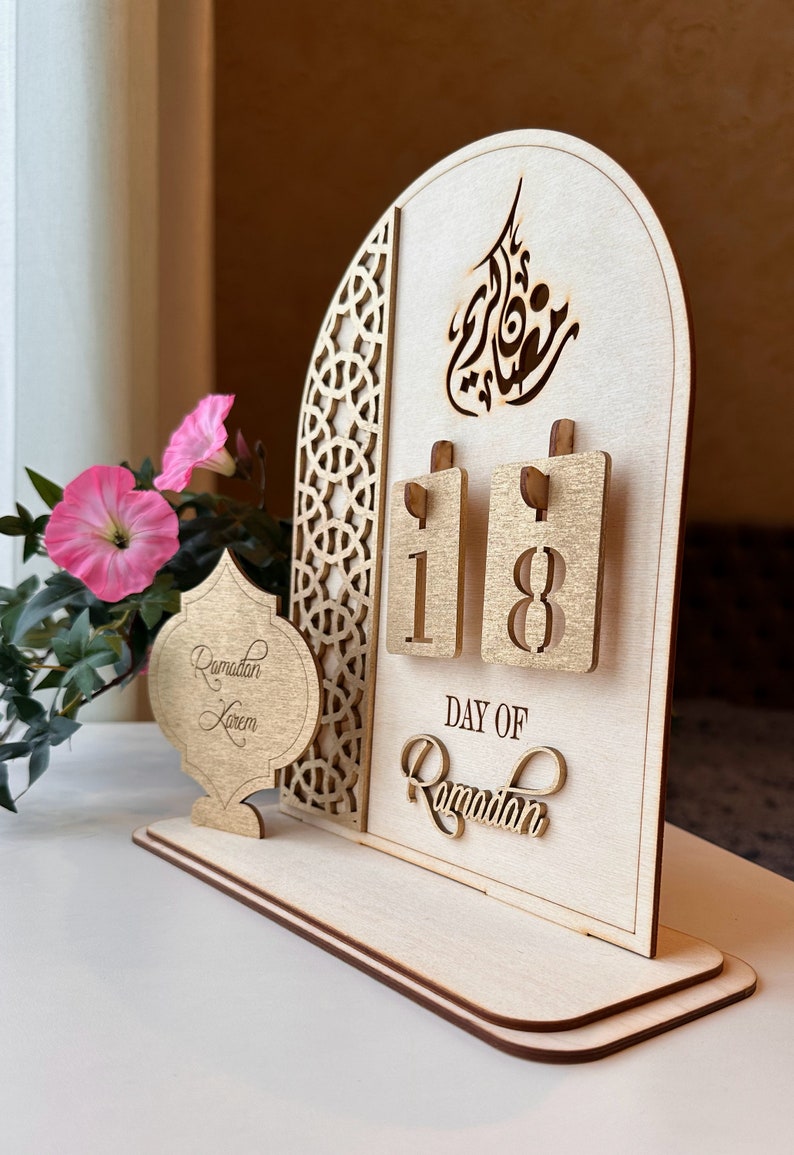 May include: A wooden Ramadan calendar with a gold-painted finish. The calendar features a decorative arch with Arabic calligraphy and a flip-style design to display the day of Ramadan. The calendar is on a wooden base and has a small wooden ornament that says "Ramadan Kareem".