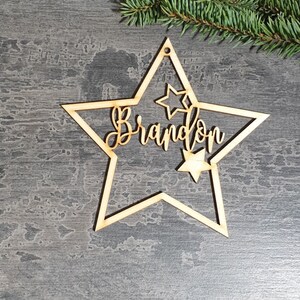 Puede incluir: Adorno navide&ntilde;o en forma de estrella de madera con el nombre "Brandon" recortado en escritura cursiva. El adorno tiene una estrella m&aacute;s peque&ntilde;a recortada en el interior y un agujero para colgarlo. Una ramita de hoja perenne est&aacute; en el fondo.