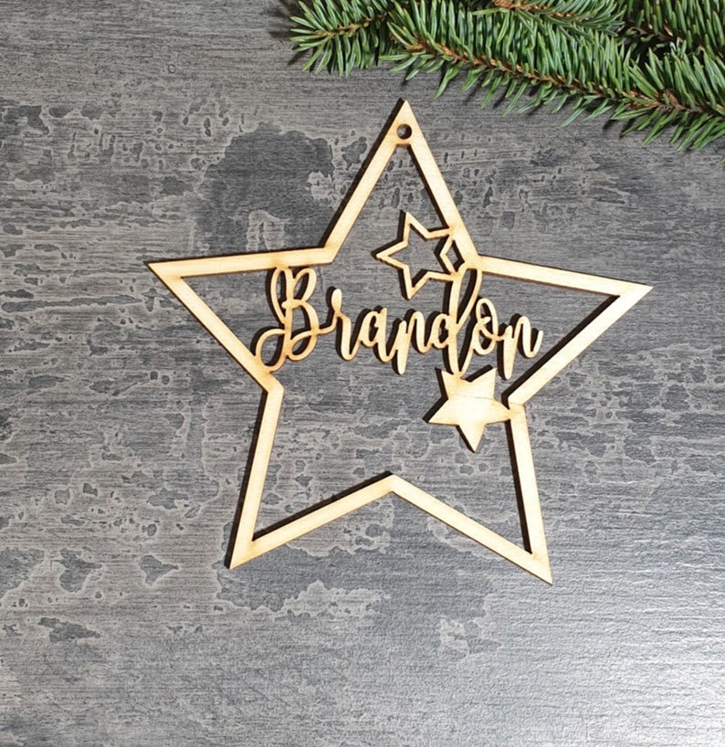 Puede incluir: Adorno navide&ntilde;o en forma de estrella de madera con el nombre "Brandon" recortado en escritura cursiva. El adorno tiene una estrella m&aacute;s peque&ntilde;a recortada en el interior y un agujero para colgarlo. Una ramita de hoja perenne est&aacute; en el fondo.