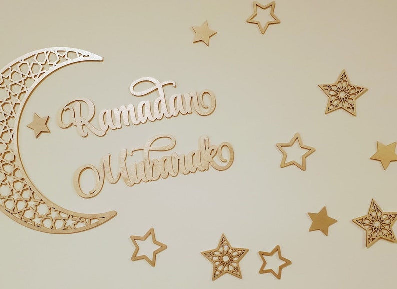 May include: A wooden crescent moon with intricate cutouts and the words "Ramadan Mubarak" in a decorative script, surrounded by gold stars of various sizes and designs.
