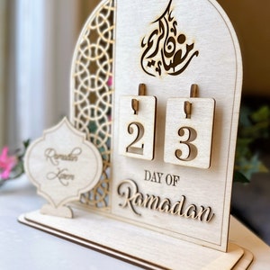 May include: A wooden Ramadan calendar with a cutout design. The calendar has two flippable numbers, currently showing 2 and 3. The text "Day of Ramadan" is written below the numbers.