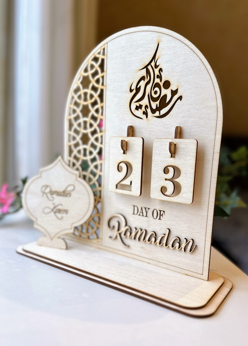 May include: A wooden Ramadan calendar with a cutout design. The calendar has two flippable numbers, currently showing 2 and 3. The text "Day of Ramadan" is written below the numbers.