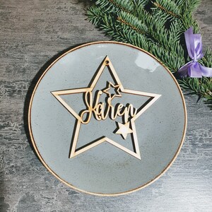 Puede incluir: Un adorno de madera en forma de estrella con el nombre "Karen" cortado en escritura cursiva. El adorno est&aacute; colocado sobre un plato gris con borde dorado, con una ramita de hoja perenne y una cinta morada en el fondo.
